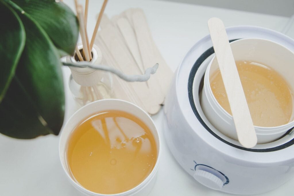 A white wax warmer contains a pot of melted, amber-coloured depilatory wax, with a wooden spatula resting inside.