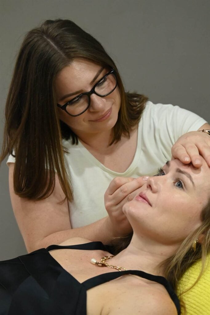 A beauty professional, wearing glasses and a white shirt, attentively working on the eyelashes or eyebrows of a blonde female client.