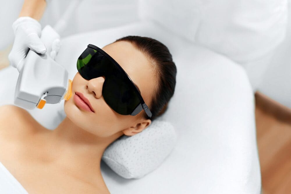 A young woman lying on a white treatment bed, wearing large black protective eyewear for a laser treatment.