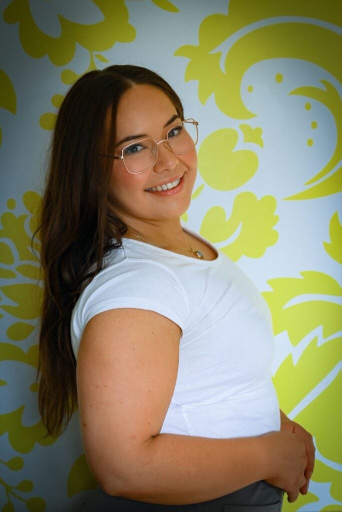 A young woman with long dark hair, wearing glasses and a white t-shirt, is smiling at the camera.