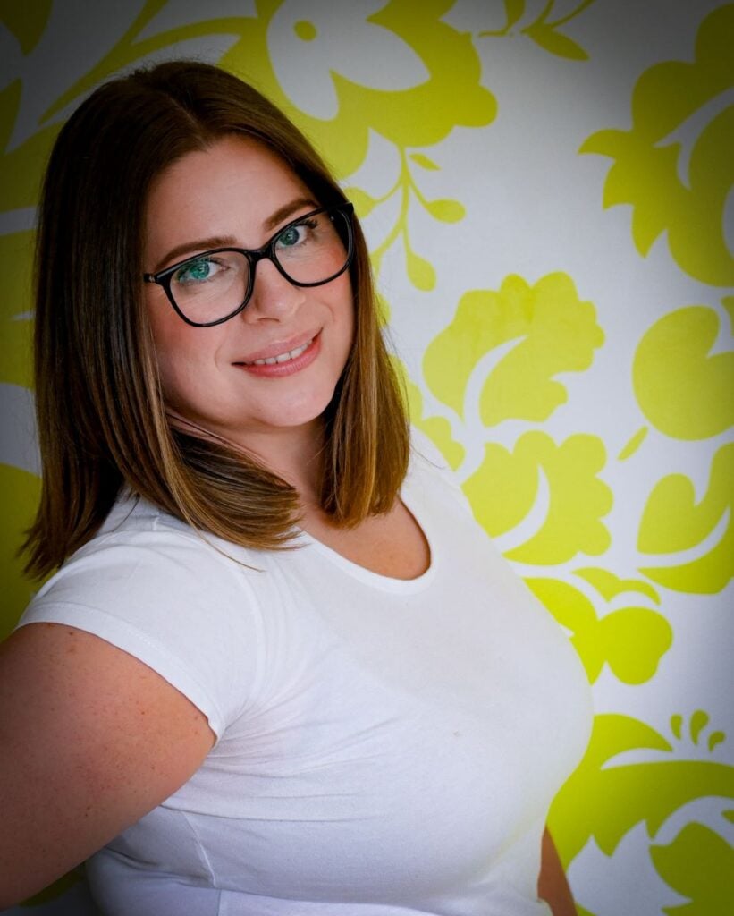 A smiling woman with shoulder-length brown hair and glasses, wearing a white t-shirt, posing indoors against a white wall with a bright yellow floral or botanical pattern.