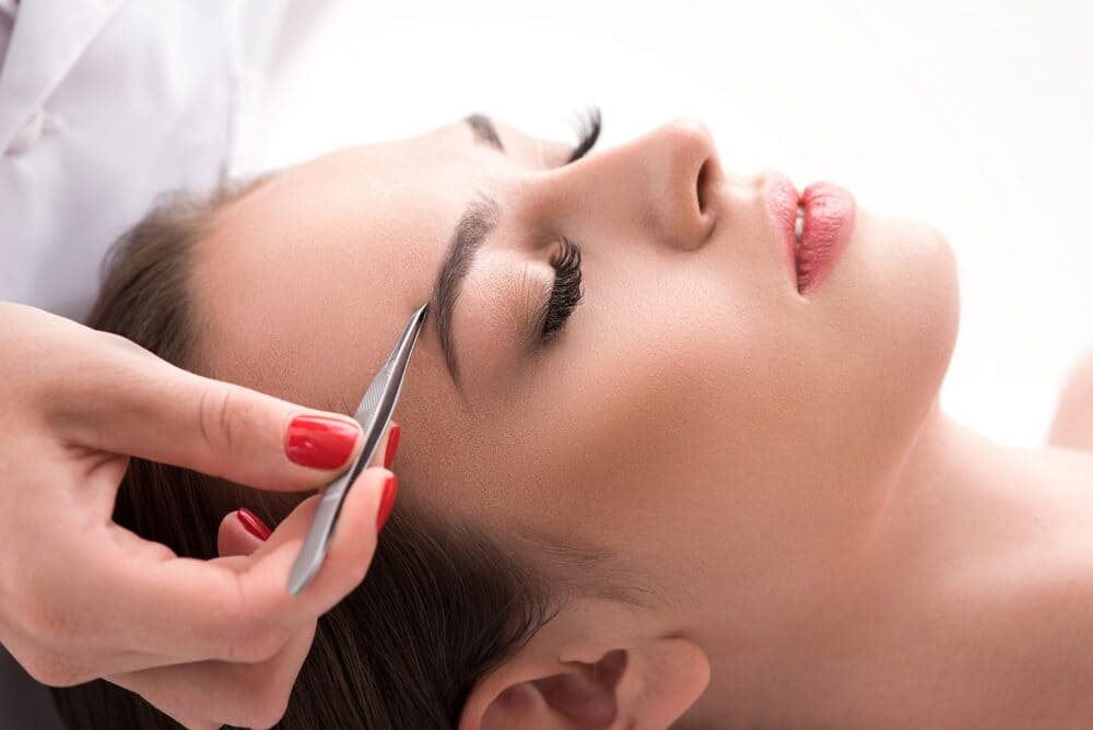 A woman with her eyes closed, lying down while receiving an eyebrow shaping treatment.