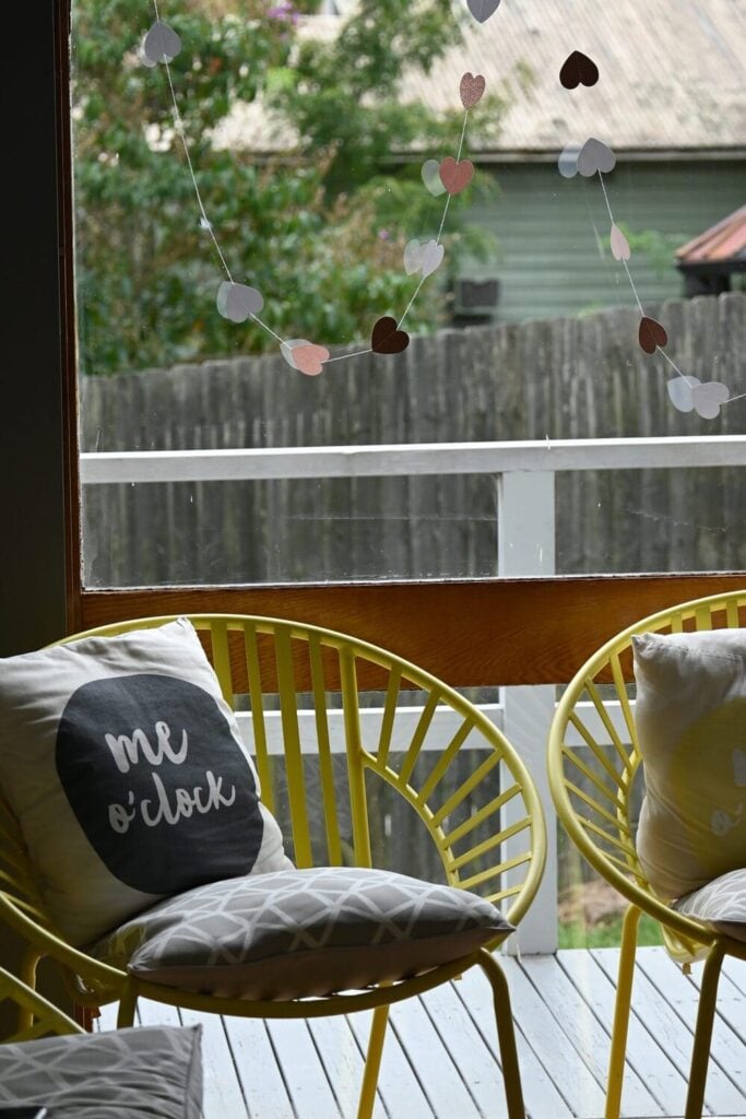 Two bright yellow wire outdoor chairs, each holding decorative throw pillows, seen through a glass door or large window.