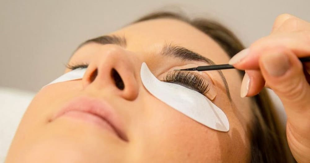 A woman lying down with her eyes closed, receiving an eyelash extension or lash lift treatment.