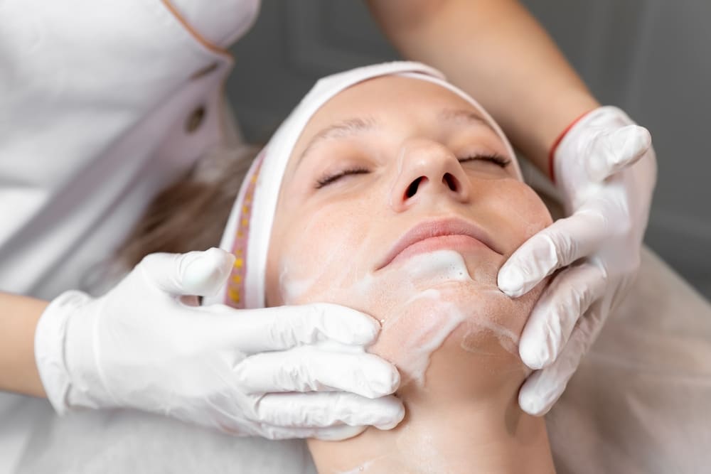 A close-up of a person lying on a treatment bed with eyes closed, receiving a facial cleansing.