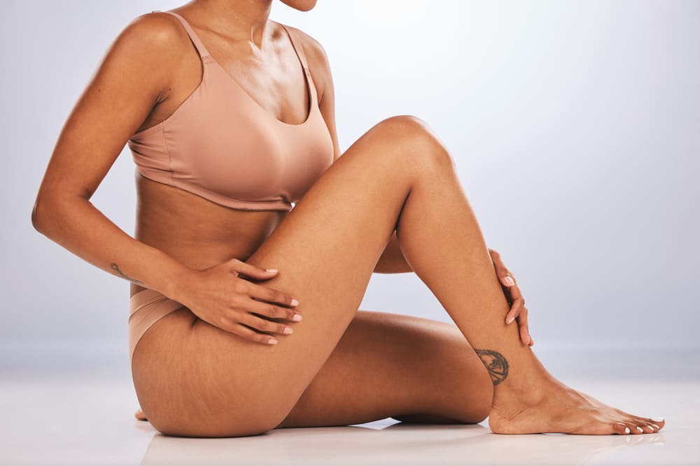 A person with medium-dark skin tone, wearing nude-colored bra and underwear, sitting on a glossy white floor against a light gray background.