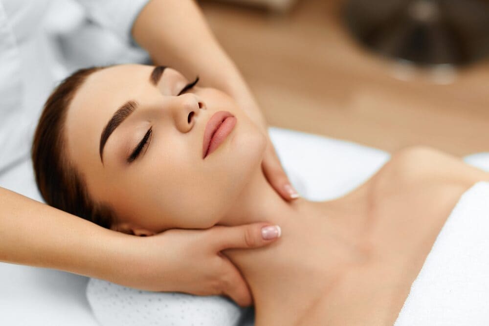 A close-up of a young woman with her eyes closed lying on a white treatment bed, looking peaceful.