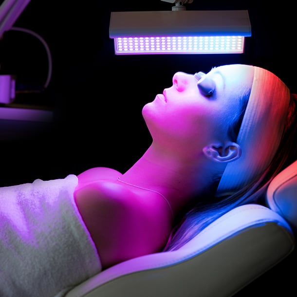 A woman lying down on a treatment bed in a darkened room, receiving an LED light therapy facial.