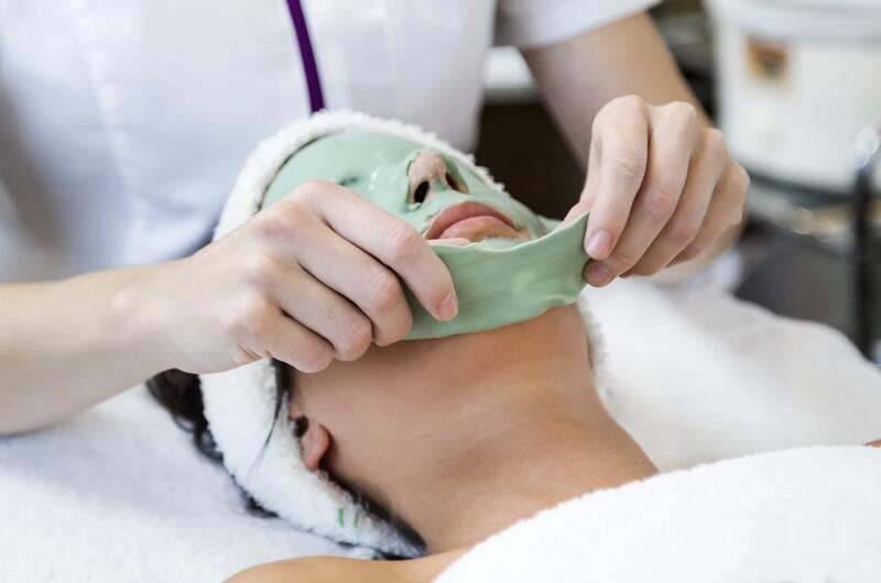 A facial treatment, while an esthetician is carefully peeling off a thick, rubbery green or mint-colored peel-off mask from the client's lower face.