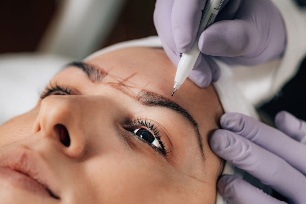 A client lying down with one eye slightly open, receiving a semi-permanent eyebrow procedure like microblading or tattooing.
