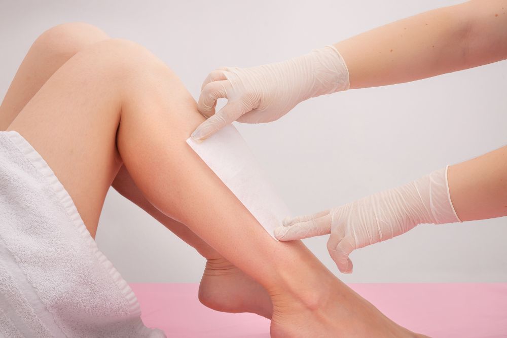 A client's smooth, bare legs lying on a surface covered with a white towel and a pink sheet, receiving a professional waxing treatment.