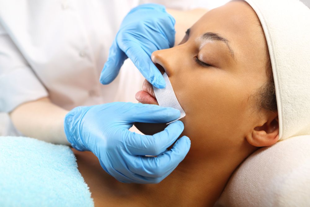 A woman lying down with her eyes closed, receiving an upper lip waxing treatment.
